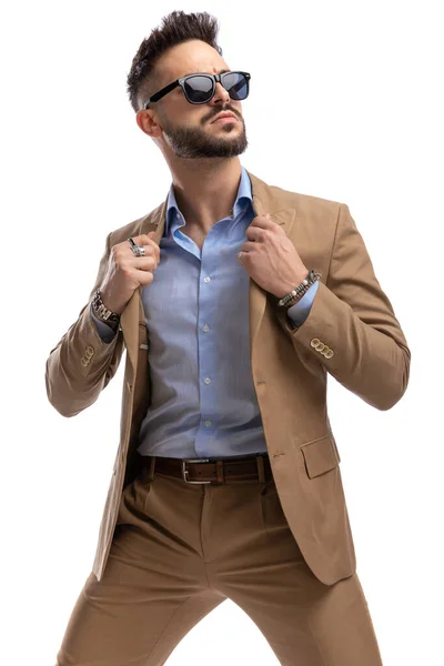 スタジオで白い背景を見ながらサングラスを引っ張ったりスーツをアレンジするクールなファッションの男 — ストック写真