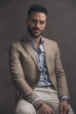 Tıraşsız, gözlüklü, bej takım elbiseli, gülen ve kahverengi arka planda, tahta koltukta oturan kendine güvensiz bir adam.