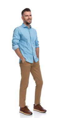 Sakallı, orta yaşlı, sıradan kıyafetli, gülümseyen ve ceplerinde el ele tutuşan bir adamın stüdyo ortamındaki beyaz arka plana bakarken tam vücut fotoğrafı.