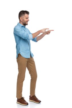 Yandan bakan, gülümseyen ve parmaklarını beyaz arka planda gösteren, mavi kot pantolonlu ve spor ayakkabılı orta yaşlı bir adamın yan görünüşü 