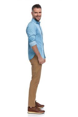 Stüdyoda beyaz arka planda beklerken gülümseyen beyaz pantolonlu ve spor ayakkabılı mutlu sakallı adamın yan görüntüsü.