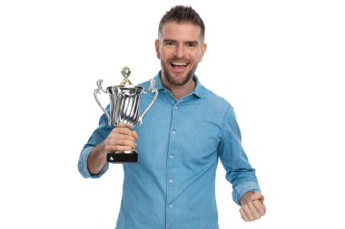 Heyecanlı sakallı adam elinde kupa tutuyor zaferi kutluyor, stüdyoda beyaz arka planda gülüyor ve gülümsüyor.