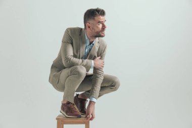 Seksi sakallı adam, dirseği dizinde, tahta koltukta çömelmiş gri arka planda, yan yatmaya çalışıyor.