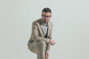 Gözlüklü, takım elbiseli, yakalı, dirseğini dizine dayayıp gri arka planda poz veren yakışıklı bir işadamı.