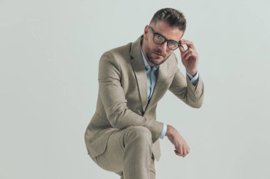 ciddi bir adam dirseğini dizine dayamış gözlük takıyor ve gri arka planda poz veriyor.