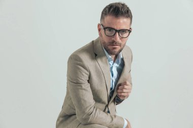 40 'lı yaşlarda, gözlüklü, dirseğini dizine dayamış, takım elbisesini ayarlayan ve stüdyoda gri arka planda kendine güvenen çekici bir adam.