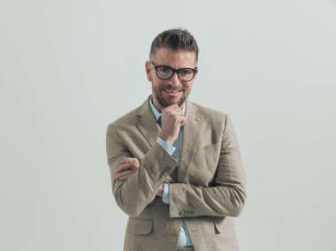 Gülümseyen, sakallı, gözlüklü, çenesine dokunan, düşünceli, hayal kuran ve gülümseyen, gri arka planda kollarını kavuşturmuş genç bir adam.