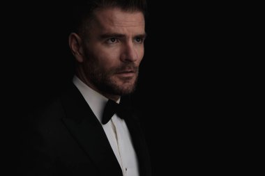 Smokin giymiş çekici bir damadın portresi yan yana duruyor ve stüdyoda siyah arka planda gelinini bekliyor.