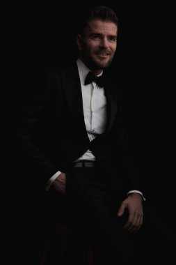 mutlu bir damat, cebinde siyah şık bir smokin giyer ve siyah arka planda otururken gelini için uzaklara bakar.