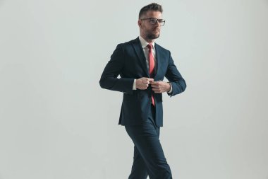 Gri arka planda stüdyoda tek başına yürürken şık takım elbiseli, kendine güvenen sakallı bir adam.