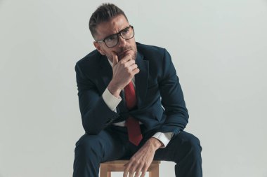 Kırklı yaşlarında, çenesine dokunan gözlüklü, kafasını kaldırıp, gri arka plandaki sorunları çözmenin yollarını düşünen bir adam.