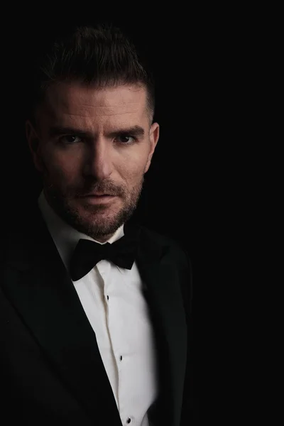 portrait of elegant good looking man in tuxedo in front of black background being mysterious in a fashion light