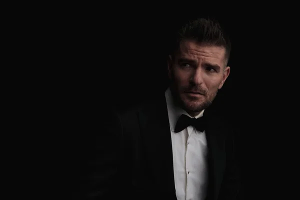 portrait of handsome bearded man in black elegant tuxedo looking up side and posing in front of black background in studio