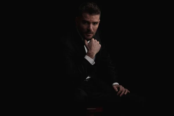 sexy businessman in black tuxedo holding hand to chin and thinking while sitting in front of black background in studio