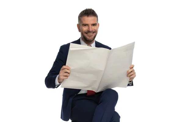 在演播室里 穿着西服的快乐而优雅的男人坐在白色背景前微笑着看报纸 双腿交叉地坐在一起 — 图库照片