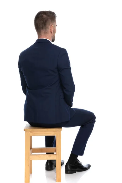 严肃的商人身穿雅致的海军蓝西服 坐在木制椅子上 背对着工作室里的白色背景 — 图库照片