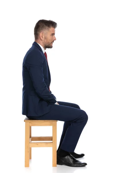スタジオの白い背景の前の木製の椅子に座ってスーツを着たエレガントな髭のビジネスマンの側面図 — ストック写真