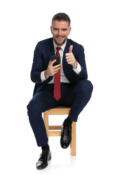 优雅的商人 穿着西服 脚背上挂着电话 坐在白色背景的木椅上 竖起大拇指 — 图库照片