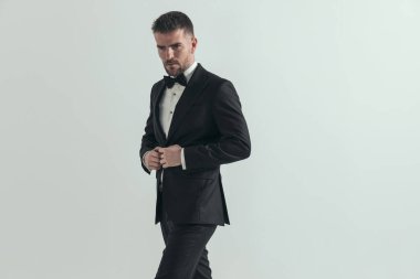 Çekici iş adamı sert bir tavırla bakışlarını kaçırıyor ve gri arka planda smokinini ayarlıyor.