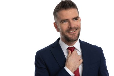 Çekici iş adamı kravatını düzeltiyor, başka tarafa bakıyor ve beyaz arka plana gülümsüyor.