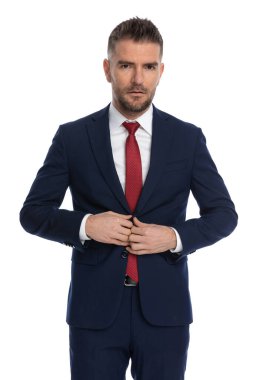 Yakışıklı iş adamı ciddi bir yüzle kameraya bakıyor ve beyaz arka planda takım elbisesini kapatıyor.