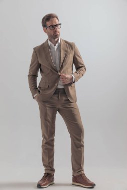 Ceketini ayarlayan, elini cebine atan ve gri arka planda poz veren çekici bir iş adamı.