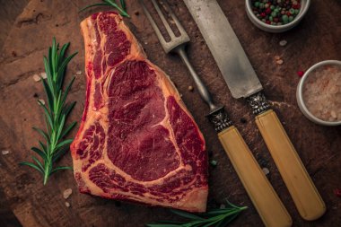 Eski ahşap kesim tahtasının üzerinde, kaliteli çatal bıçak takımı, biberiye, pembe tuz ve renkli biber kaseleri olan biftek estetiğinin en üst görüntüsü. 