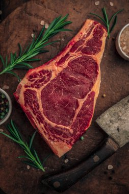 Eski paslı kesme tahtasının üzerinde biftek, bitkiler, küçük kase renkli biber ve yanında büyük bir bıçak ve tuz serpiştirilmiş pembe tuz. 