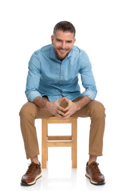 Stüdyoda beyaz arka planda otururken dirseklerini dizlerinin üstüne çöküp gülümseyen orta yaşlı bir adam.