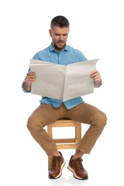 Kot gömlekli meraklı adam beyaz arka planda stüdyoda otururken gazete okuyor.