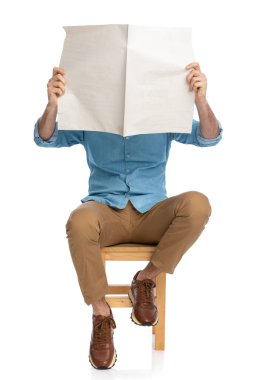 Spor ayakkabılı, spor ayakkabılı, gazete yüzlü sıradan bir adam. Stüdyoda beyaz arka planda, tahta bir sandalyede oturuyor ve kitap okuyor.