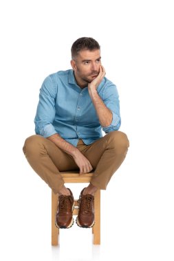 Kot gömlekli, düşünceli, orta yaşlı bir adam. Sandalyeye oturmuş stüdyoda beyaz arka planda poz verirken düşünüyor.