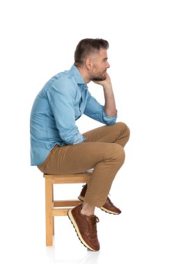 sexy thoughtful man in denim shirt looking to side and thinking while posing on wooden chair with elbow on knee on white background