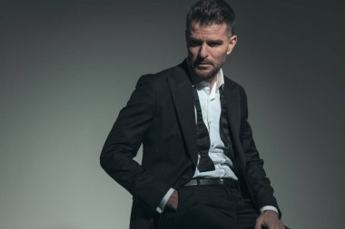 Smokinli, çözülmüş gömlekli, seksi ve zarif bir adam gözlerini kaçırıp gri arka planda el ele poz veriyor.