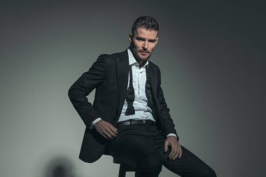 Tıraşsız orta yaşlı seksi adam gri arka planda otururken kendine güvenen bir şekilde poz veriyor. 