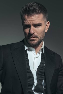 Kendine güvenen, zarif takım elbiseli, çözülmemiş kravatlı, gizemli ve gri arka planda poz veren seksi adam.