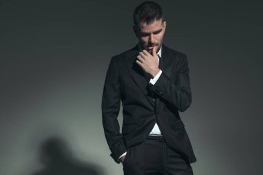 handsome middle aged man in elegant tuxedo sensually touching lips and looking down while posing in a cool manner on grey background