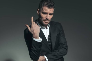 Şık smokinli seksi sağdıç moda ışığında poz verirken gri arka planda kendine güvenen bir şekilde parmağını kaldırıyor.