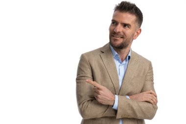 Tıraşsız iş adamının bej takım elbiseli portresi. Gömleği çıkarılmış, sağa sola bakıyor, gülümsüyor ve stüdyoda beyaz arkaplan önünde poz veriyor.