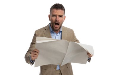 astounded businessman in beige suit with undone shirt opening mouth and reading newspaper on white background in studio