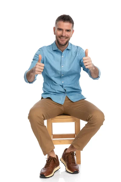 坐在身穿蓝色牛仔裤衬衫和奇诺裤子的四十多岁的快乐而随意的男人面前 坐在白色背景的木椅上做手势微笑 — 图库照片