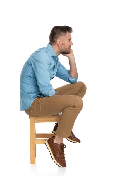 白い背景に膝の上に肘で木製の椅子に身を置きながら脇と思考を探しているデニムシャツのセクシーな思慮深い男 — ストック写真