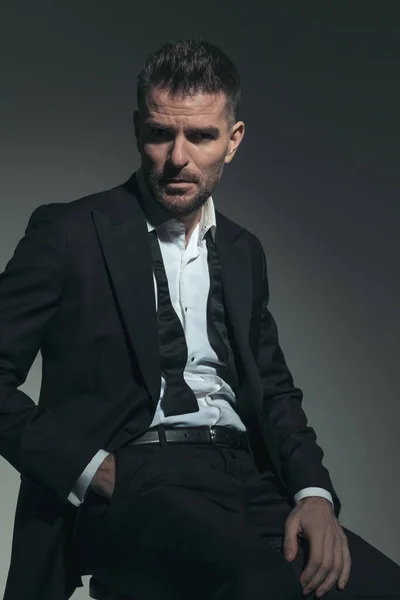 dramatic businessman in tuxedo with undone tie being mysterious and posing with hand in pocket while sitting on grey background