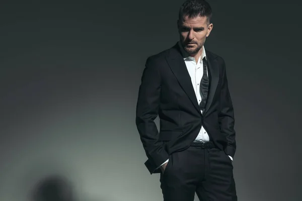 sexy confident man in tuxedo holding hands in pockets and looking to side on grey background in studio