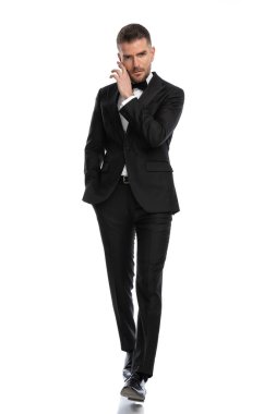 Çekici iş adamı kameraya doğru yürürken yüzüne dokunuyor ve bir elini cebinde tutuyor.