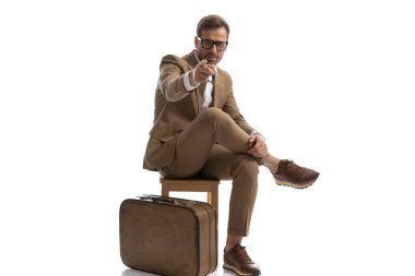 Genç yakışıklı iş adamı kamerayı işaret ediyor, bacağını tutuyor ve bavulunun yanında oturuyor.