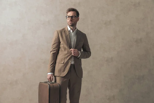 cool businessman arranging his jacket while looking to side and holding a luggage against wallpaper