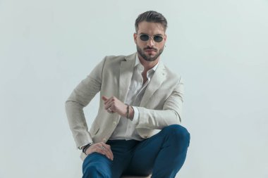 Dizlerine dirsek atıp poz veren, güneş gözlüğü takan ve stüdyoda gri arka planda kendine güvenen havalı iş adamının portresi.