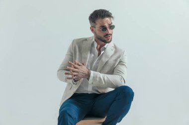 Kendine güvenen iş adamının portresi, eski güneş gözlükleriyle avuçlarına sürterken, yan tarafa bakarken ve gri arka planda tahta koltukta otururken. 