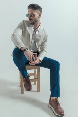 seductive businessman with undone shirt touching fingers and looking to side while sitting on wooden chair in a cool manner for posing 